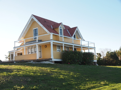 [This two-story yellow house  has porches on the first two levels. It has a bay window on the left side and large bushes which come up more than half the height of the first floor. Two windowed dormers face the water.]